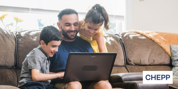 Father sitting on couch working on computer with children leaning over shoulder, all smiling