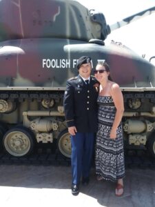 Daniel in military uniform standing and smiling with a woman in a patterned dress in front of a military tank