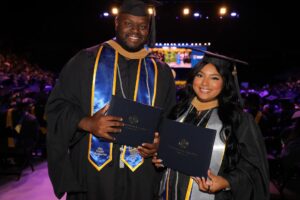 Gasten and Mandy in their regalia smiling at the camera holding their diplomas.