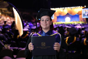 Graduate in cap and gown smiling and holding a Southern New Hampshire University diploma during a commencement ceremony.