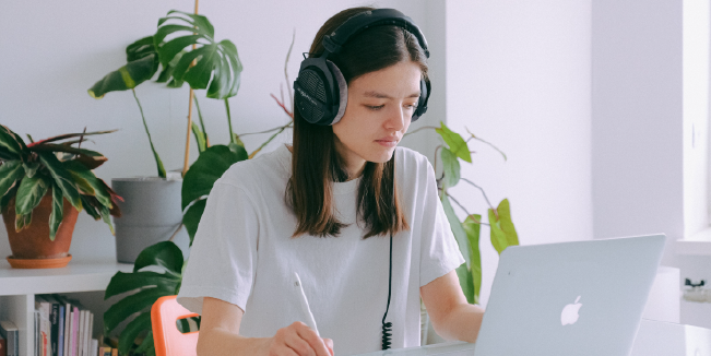 Student wearing headphones while using a laptop and taking notes at a desk in a bright study space.