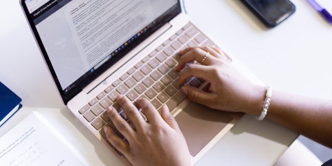 Close-up of a person typing on a laptop at a desk with a notebook, smartphone, and pen nearby.