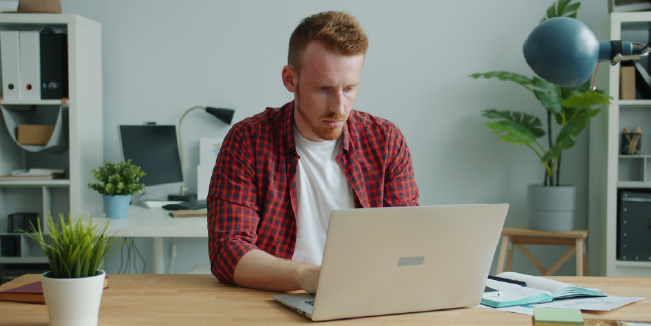 Person working on a laptop at a desk in a home office with notebooks and plants nearby.
