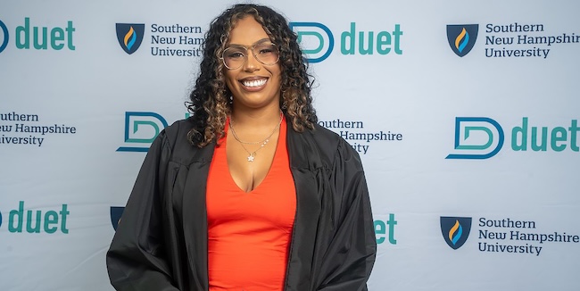 Person wearing glasses, a red top, and black graduation regalia stands in front of a backdrop featuring Southern New Hampshire University and Duet logos.